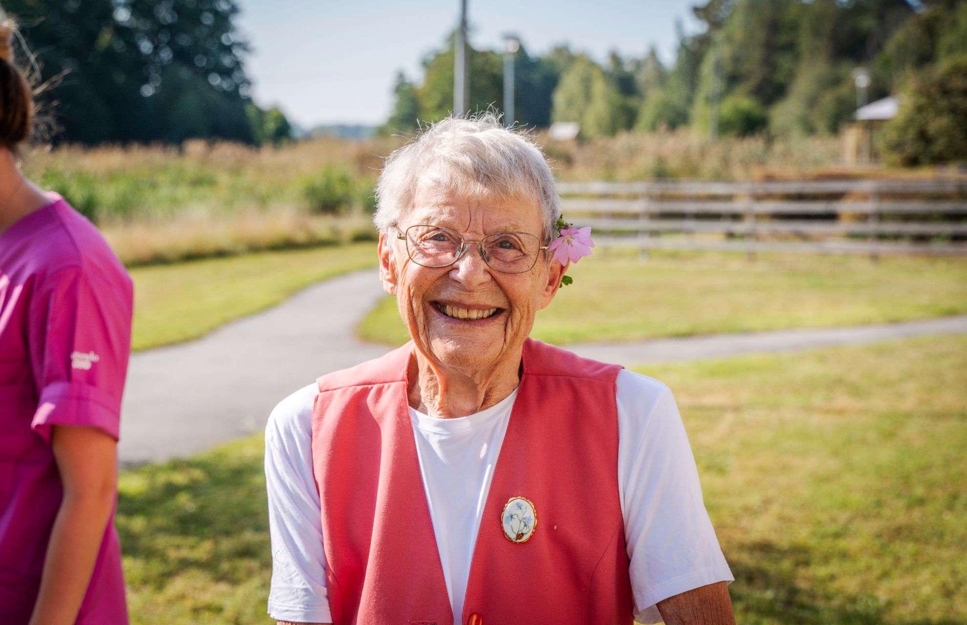 Äldre kvinna på en grön äng med en blomma bakom örat och ett stort leende