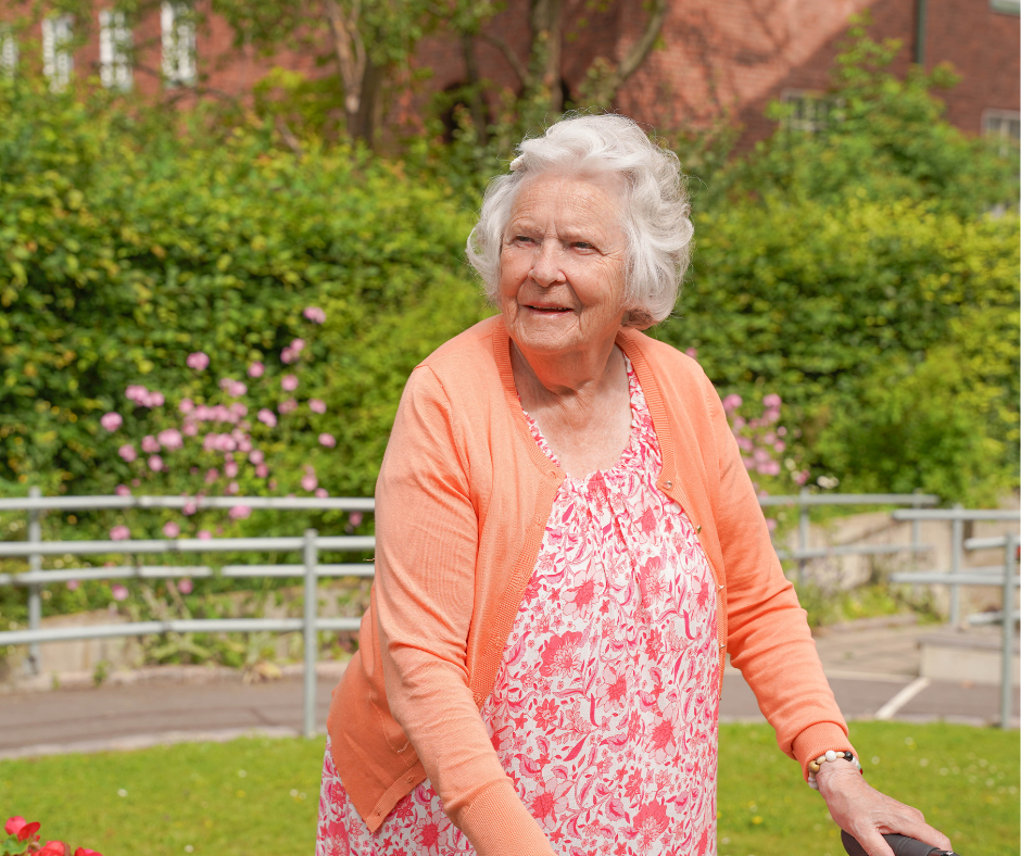 Äldre dam  i färgglada kläder står i grön omgivning och spanar ut i trädgården.
