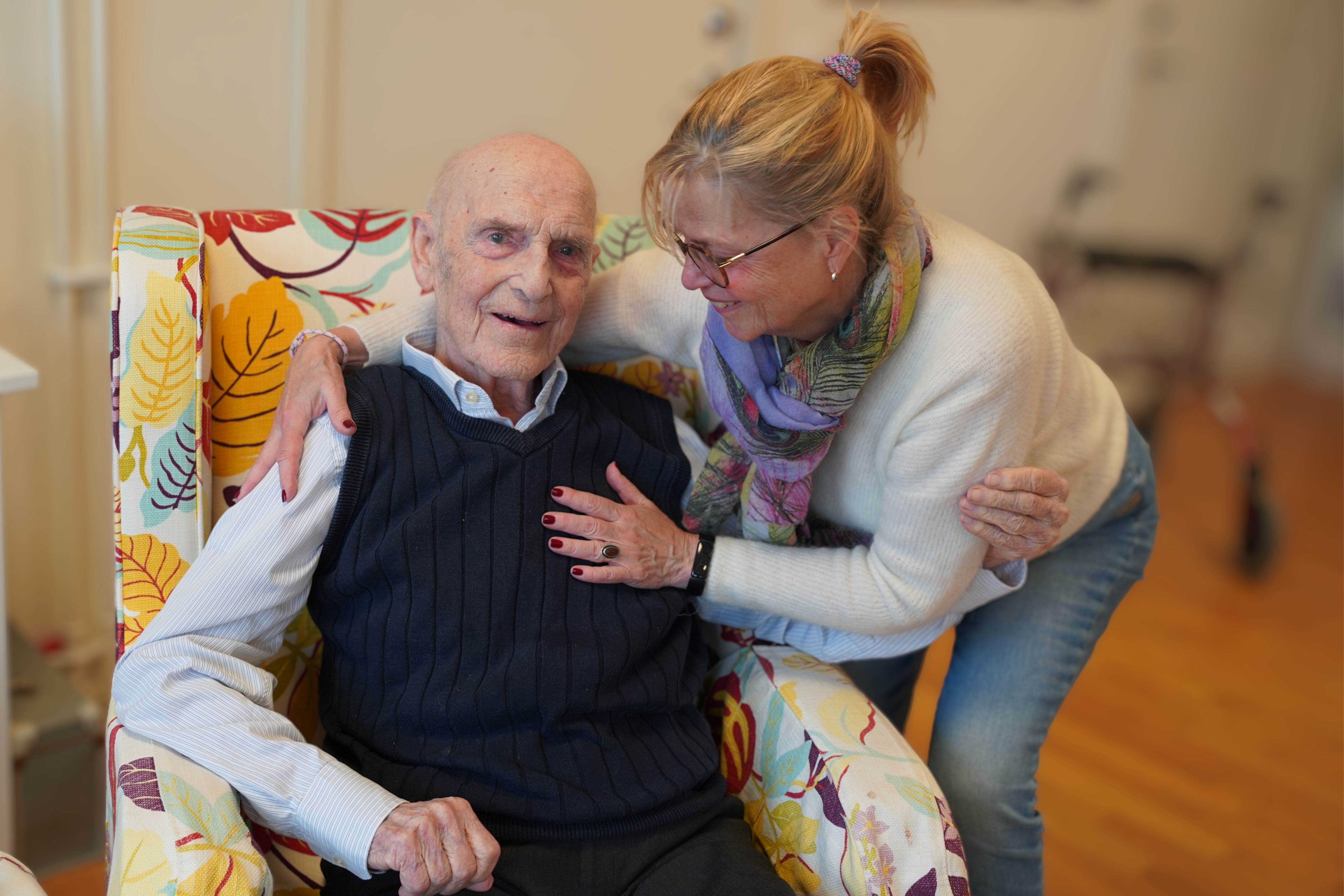 Äldre man i randig väst sitter i fåtölj med färgglatt mönster, omgiven av sin dotter, en medelålders kvinna med glasögon och halsduk i ljus tröja.